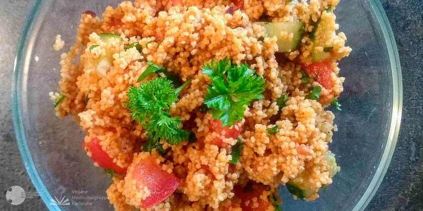 Couscous Salat, in einer Glasschüssel. Ganiert mit zwei Petersilienblättern. Im Couscous Salat sieht man Tomaten, Gurken und kleingeschnittene Petersilienblättern. Der Couscous Salat ist durch den Dressing leicht orange angehaucht.