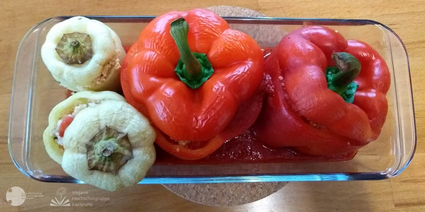 In einer gläsernen Auflaufform, sind zwei rote angeschnittene Paprika und zwei kleine weiße Paprika in eine rote Soße gesetzt.