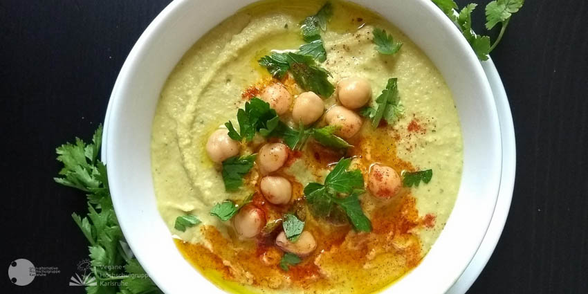 Eine Schale Humus ist mit einigen Kichererbsen, Kräutern und Gewürzen garniert. Diagonal angrenzend sind weitere Sträuschen Kräuter angeordnet, die sich vom schwarzen Tisch abheben.