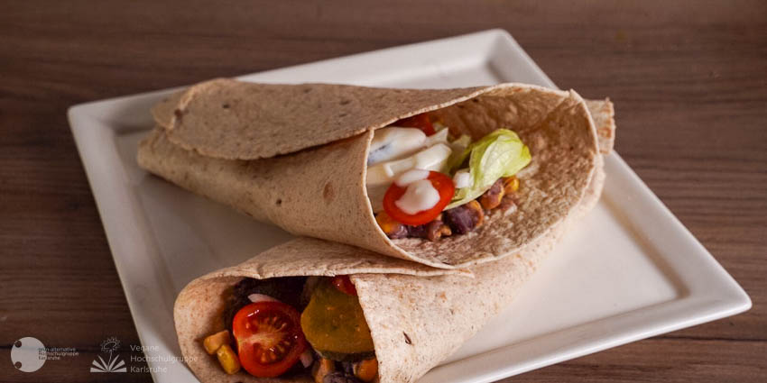 Auf einem quadratischen weißen Teller, der auf einem dunkelbraunen Holztisch steht, sind zwei Mais-Bohnen-Wraps über Kreuz angeordnet. Neben dem Mais und den Bohnen sind Tomaten, Salat, Zwiebelstreifen und Dressing als Füllung sichtbar.