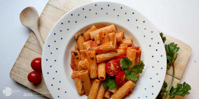 In Sauce gewälzte Nudeln liegen, garniert mit einer kleinen Tomate und Koriander, in einem weißen Teller mit blau getüpfeltem Rand. Er steht auf einem Holzbrettchen, auf dem ein hölzerner Kochlöffel, zwei kleine Tomaten und ein Stengel Koriander liegen.