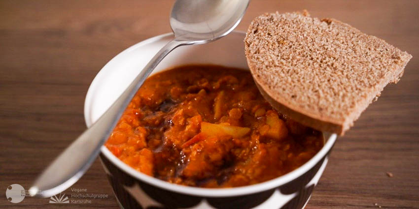 Eine auf der Außenseite mit schwarzen Kreisen verzierte weiße Schüssel ist mit Rote Linsen Dal gefüllt. Auf ihrem Rand liegen links ein Esslöffel und hinten rechts eine halbe Brotscheibe.