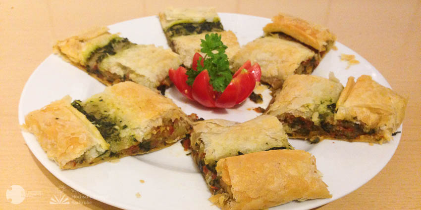 Sechs Spinatstrudel-Stücke sind sternförmig auf einem weißen Teller um eine blütenförmige Tomate angeordnet.