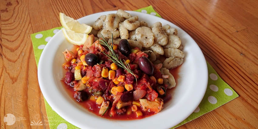 Auf einem Holztisch steht ein mit Serviette unterlegter runder Teller, der hälftig mit der rot-gelb dominierten Tomatensauce und mit Gnocci gefüllt ist. Das Gericht ist mit zwei Zitronenspalten und einem Zweig Rosmarin garniert.
